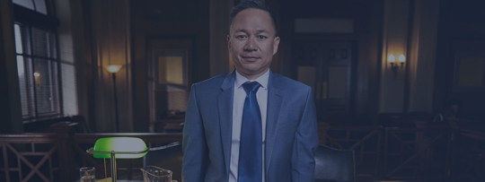 A lawyer in a suit stands in a dimly lit courtroom.